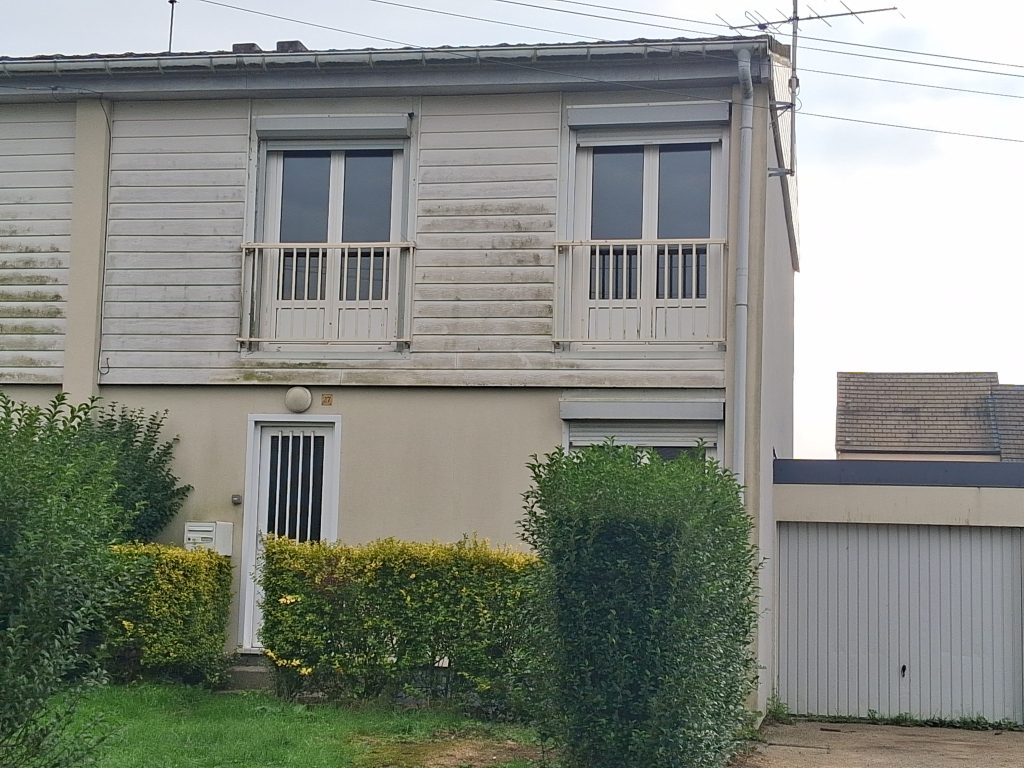Petite maison sur un étage, avec un garage blanc sur le côté à droite. En bas, on voit la porte d'entrée et une haie verte. En haut, il y a deux fenêtres avec des barrières blanches. Un petit jardin avec de l'herbe se trouve devant la maison.