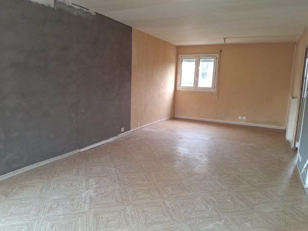 Une grande pièce vide avec un sol clair à motifs. Le mur de gauche est gris foncé et le mur du fond est beige avec une fenêtre blanche. Le plafond est blanc et plat.