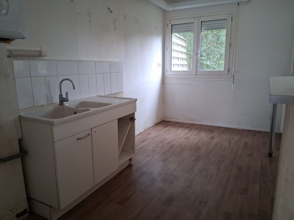 Cuisine non équipée. Le meuble vasque, situé à gauche de la photo est blanc, il dispose de deux portes, mais n'est pas équipé. Le sol est un parquet marron. Au fond de la pièce il y'a une double fenêtre blanche. Les murs sont également blancs.